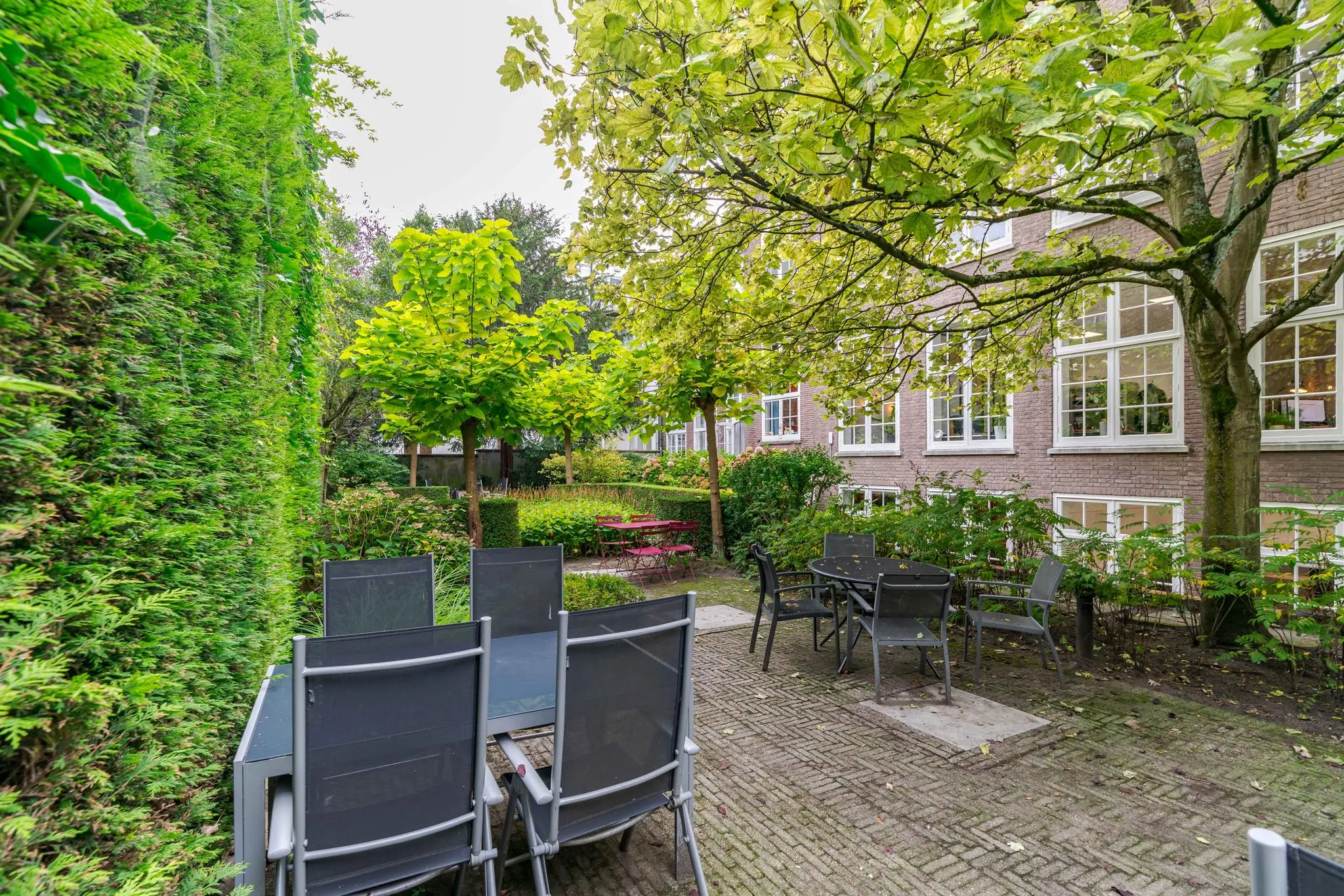 Binnentuin aan het Lange Voorhout met terrasmeubilair, groene hagen en grote bomen grenzend aan een gebouw met witte raamkozijnen.